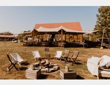 Eine outdoor Szene mit einer Sitzgruppe aus Holz- und Liegestühlen, die um eine Feuerstelle angeordnet ist. Im Hintergrund steht ein großes Zelt mit Holzstühlen und Tischen, das vor einem Gebäude mit rotem Satteldach und Holzwänden aufgestellt ist.