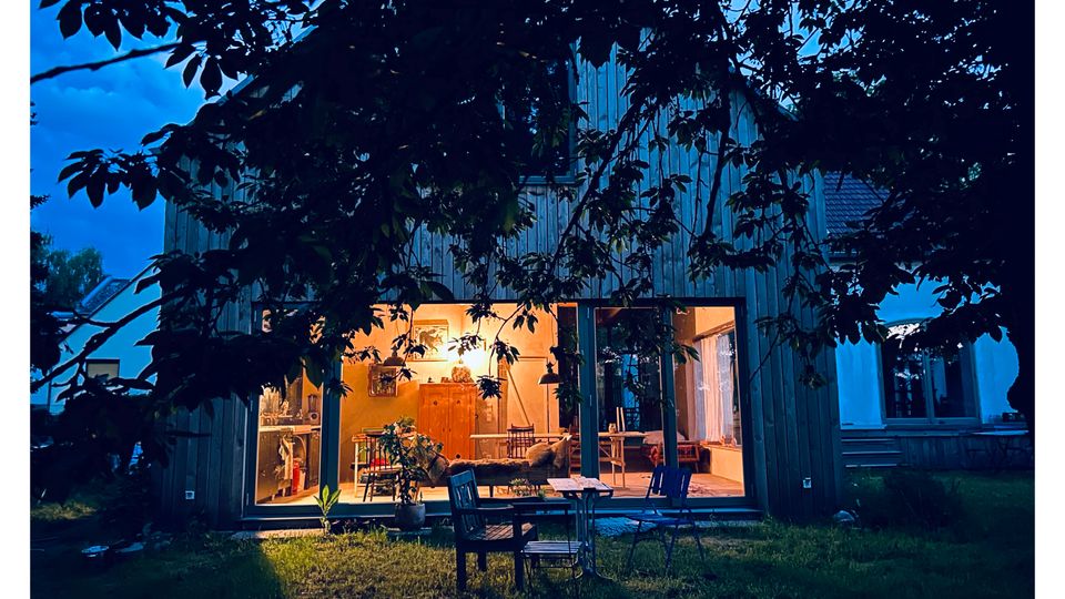 Holzverkleidetes Wohnhaus in der Dämmerung mit großem, bodentiefem Glasfenster und warm beleuchtetem Innenraum. Davor liegt ein Rasen mit Terrasse und Gartenmöbeln, teilweise von Baumästen im Vordergrund eingerahmt.