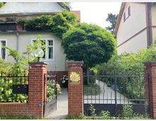 Zweistöckiges Wohnhaus mit hellem Putz und Ziegeldach, umgeben von dichtem Grün; ein Zierbaum und Rankenpflanzen schmücken den Eingangsbereich. Im Vordergrund befindet sich ein schmiedeeisernes Tor mit Backsteinpfeilern, hinter dem ein gepflasterter Weg zum Haus führt.