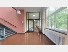 Ein Schulflur mit glänzendem braunem Boden, einer Treppe auf der linken Seite, großen Fenstern auf der rechten Seite, die Blick auf grüne Bäume bieten, und einer Glastür, die zu einem langen, hellen Gang führt.