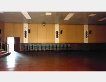 Leerer Raum mit braunem Holzboden und holzvertäfelten Wänden, an denen mehrere Stühle in einer Reihe aufgestellt sind. An den Wänden befinden sich Lautsprecher und Beleuchtungselemente, und links ist eine geöffnete Tür sichtbar.