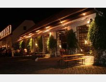 Nachtaufnahme eines Restaurants mit beleuchtetem Außenbereich. Die Fassade des Gebäudes ist mit großen Glasfenstern und Rankenpflanzen verziert, davor stehen Bierbänke und Tische auf einem Steinpflaster.