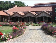 Ein gepflasterter Weg führt durch einen gepflegten Garten mit bunten Blumenbeeten, umgeben von einer Anlage mit Holz- und Ziegelarchitektur. Im Hintergrund sind Stallgebäude mit roten Ziegeldächern und bogenförmigen Eingängen zu sehen.