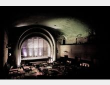 Dunkler Saal mit einer bogenförmigen Bühne im Hintergrund, akzentuiert durch violette Beleuchtung. Im Vordergrund sind mehrere verstreute Tische und Stühle sichtbar, die eine Art Veranstaltungs- oder Konzertort andeuten.