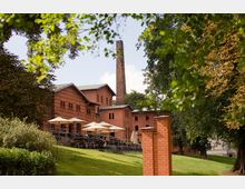 Backsteingebäude mit einem hohen Schornstein in einer grünen Parklandschaft, umgeben von Bäumen. Im Vordergrund befindet sich eine Terrasse mit Tischen und Sonnenschirmen.