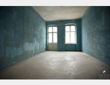 Ein leerer, rechteckiger Raum mit abgenutzten, türkisfarbenen Wänden, einem hellen, beigen Boden und einer hohen Decke. An der Stirnwand befinden sich zwei große Fenster mit Rundbögen und eine Tür mit Glaseinsätzen, durch die Tageslicht einfällt.