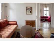 Wohnzimmer in einem Altbau mit Parkettboden, einem orangefarbenen Sofa, einem ovalen Holztisch und einer antiken Holzkommode. Im Hintergrund ist ein weiterer Raum mit roten Polstermöbeln und großen Fenstern sichtbar, die viel Tageslicht einlassen.