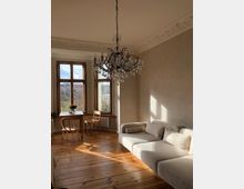 Helles Wohnzimmer mit Holzboden, einer cremefarbenen Eckcouch und einem prunkvollen Kronleuchter. Große Sprossenfenster bieten Blick auf eine Landschaft, und ein kleiner Tisch mit zwei Stühlen steht vor dem Fenster.