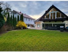 Zwei Einfamilienhäuser in einer grünen Gartenlandschaft, das vordere Haus mit großer Fensterfront und Holzgiebel. Im Garten sind Sträucher, Koniferen sowie eine Terrasse mit Sitzmöbeln sichtbar.