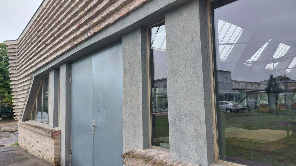 Gebäude mit moderner Stein- und Putzfassade, großen Fenstern und einer Metalltür; im Hintergrund sind Glasdach-Reflexionen, parkende Autos und Grünflächen erkennbar.