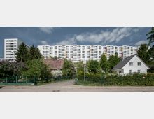 Straßenszene mit einem Kontrast zwischen kleinen Einfamilienhäusern mit Gärten im Vordergrund und einer langen Reihe weißer Wohnhochhäuser mit Balkonen im Hintergrund; umgeben von Bäumen und Grünflächen unter einem bewölkten Himmel.