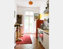 Schmale Küche mit Terrakotta-Fliesenboden, rotem Teppich und weißen Wänden. Die Einrichtung umfasst einen roten Kühlschrank, eine helle Holztheke mit Hockern, ein modernes Cerankochfeld und ein Fenster mit Doppeltür, das auf einen kleinen Balkon führt.