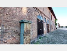 Backsteinfassade eines historischen Gebäudes mit dunklen Holztüren und Fenstern mit Bögen. Im Vordergrund steht ein verwitterter, metallener Schaltkasten; der Boden besteht aus unebenem Kopfsteinpflaster.