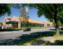 Eine Backstein-Gebäudestruktur mit orangefarbenem Satteldach und großen Fenstern, die an einer ruhigen Straße mit Bäumen steht. Im Vordergrund ist ein geparkter schwarzer Wagen und ein umzäuntes Gelände zu sehen.