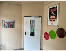 Innenbereich eines Diners oder Cafés mit beigen Wänden und einer weißen Tür mit schmalem Sichtfenster in der Mitte. An den Wänden hängen ein Burger-Werbeplakat, ein Foto sowie drei runde, farbige Tafeln mit handschriftlichen Notizen.