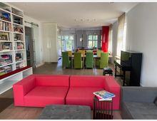 Großzügiges Wohnzimmer mit offener Verbindung zur Küche. Im Vordergrund stehen rote und graue Sofas, rechts ein schwarzes Klavier, links ein Bücherregal, hinten ein Esstisch mit grünen Stühlen und im Hintergrund die moderne Küche mit roten Akzenten.