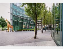 Moderner Gebäudekomplex mit einer gläsernen Fassade, umgeben von gepflasterten Gehwegen und Bäumen; im Vordergrund steht eine große Giraffenfigur, und im Hintergrund ist ein buntes Eingangsbereich-Design sichtbar.