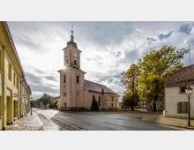 Straßenszene einer Kleinstadt mit einer hellen Kirche im Barockstil, die einen hohen Turm mit Uhr besitzt. Links der Kirche stehen Wohnhäuser, rechts befinden sich Bäume und weitere Gebäude; der nasse Asphalt reflektiert das Licht eines bedeckten Himmels.