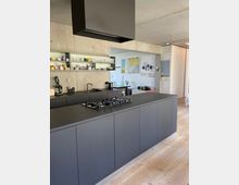 Moderne Küche mit einer großen, mattschwarzen Kücheninsel mit integriertem Gaskochfeld und einer schlichten Arbeitsplatte. Die Rückwand aus Sichtbeton enthält offene Regale mit Geschirr, und der Boden besteht aus hellem Holz.