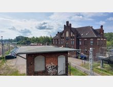 Alter Bahnhofsbereich mit einem Backsteingebäude im historischen Stil, flankiert von Gleisanlagen und einem kleinen Parkplatz. Im Vordergrund ein Nebenbau mit Graffiti, dahinter Waldbestand und vereinzelte Züge auf den Gleisen.