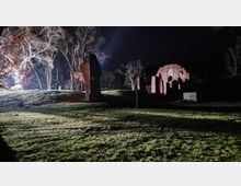 Nachtaufnahme einer Ruine aus rotem Backstein, umgeben von Bäumen und beleuchtet durch künstliches Licht. Im Vordergrund ist eine grasbewachsene Fläche zu sehen, während im Hintergrund die Struktur der Ruine mit Bögen und Mauerresten erkennbar ist.