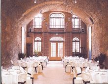 Großer Raum mit einer gewölbten Backsteindecke und großen Fenstern, die vom Boden bis zur Decke reichen. Der Raum ist für eine Veranstaltung vorbereitet, mit runden Tischen, weiß eingedeckten Stühlen und dezenter Beleuchtung.