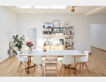 Helles Esszimmer mit weißem Tisch, umgeben von sechs Stühlen, auf einem Holzboden. Im Hintergrund stehen ein Bücherregal, ein großer Spiegel, Zimmerpflanzen und Dekorationsobjekte; von oben fällt Tageslicht durch ein Oberlicht ein.