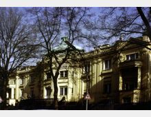 Gelbes, historisches Gebäude mit klassizistischen Elementen wie Säulen und Verzierungen, umgeben von großen, unbelaubten Bäumen. Im Vordergrund sind ein schmiedeeiserner Zaun und ein Verkehrsschild für einen Fußgängerübergang sichtbar.