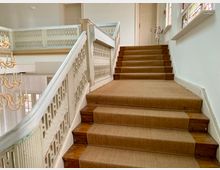 Treppenaufgang in einem historischen Gebäude mit einem Geländer aus weiß lackiertem Holz und dekorativen Verzierungen. Die Treppenstufen sind mit einem hellbraunen Teppich bedeckt, seitlich sind helle Wände mit einem Kerzenständer und bunten Fenstern sichtbar.