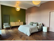 Modern bedroom with a low upholstered bed dressed in neutral linens, set on a dark herringbone wood floor. The room features a green accent wall with a radiator and wooden sideboard, a gray armchair, soft pink walls, and two sculptural pendant lights.
