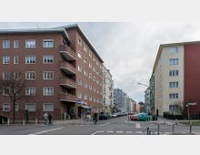Straßenecke in einer Wohngegend mit mehrstöckigen Wohngebäuden; links ein braunes Gebäude mit Balkonen, rechts ein hellgelbes Gebäude. Autos parken entlang der Straße, und mehrere Fußgänger und ein Fahrradfahrer sind unterwegs.
