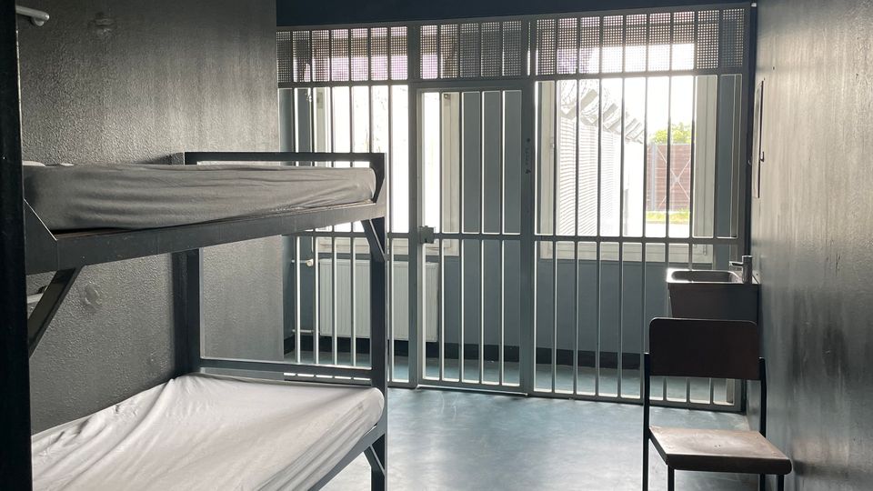 Schmale, karge Zelle mit grauen Wänden, einem Metall-Etagenbett mit dünnen Matratzen und einem einzelnen Stuhl. Am Ende des Raums befindet sich eine vergitterte Tür vor einem Fenster, durch das Tageslicht einfällt.