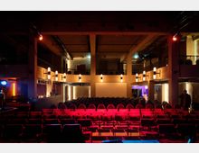 Innenansicht eines Veranstaltungsraumes mit Reihen aus roten Bestuhlungselementen, einem Bühnenbereich im Hintergrund und modernen Wandleuchten. Der Raum hat einen offenen, zweistöckigen Aufbau mit Balkonen und ist in warmes, gedämpftes Licht getaucht.