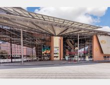 Moderne Glasfassade des Gebäudes der Spielbank Berlin am Potsdamer Platz mit markantem Vordach aus Stahl und Glas. Im Vordergrund ein gepflasterter Platz und Eingangsbereiche mit Plakaten und Beschilderungen.