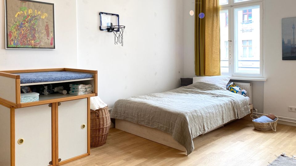 Helles Schlafzimmer mit Holzfußboden, einem Einzelbett neben einem großen Fenster mit goldenen Vorhängen und einer Kommode mit Wickelauflage. An der weißen Wand hängen ein abstraktes Bild und ein kleiner Basketballkorb; daneben stehen Körbe und ein kleines Babybettchen am Boden.