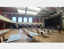 Ein großer Veranstaltungssaal mit holzfarbenem Boden, einer Bühne auf der rechten Seite und mehreren Reihen von Tischen und Stühlen, die in der Mitte des Raumes angeordnet sind. Große Fenster, über denen rote Vorhänge hängen, lassen Tageslicht herein, und die Decke ist mit Licht- und Akustikinstallationen ausgestattet.