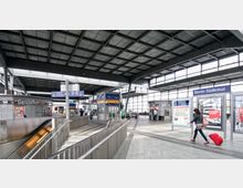 Innenansicht des Bahnhofs Berlin Südkreuz mit moderner Stahl- und Glasarchitektur. Zu sehen sind Beschilderungen, Rolltreppen, Fahrkartenautomaten und Plakate, während Menschen mit Gepäck durch die Halle gehen.