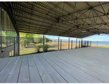 Offene, überdachte Holzplattform mit Metallgitterwänden, umgeben von einer natürlichen Landschaft mit Bäumen, Schilf und Blick auf ein Gewässer im Hintergrund. Die Dachkonstruktion besteht aus wellenförmigem Metall mit sichtbaren Streben.