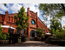 Backstein-Gebäude mit historischen Elementen und Rundfenster, umgeben von Bäumen und einer Außenterrasse mit Tischen und Stühlen; im Hintergrund ein Gerüst eines Gasometers sichtbar.