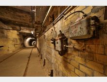 Ein alter, schmaler Tunnel mit gelben Ziegelwänden und sichtbaren Elektroinstallationen. An der rechten Wand sind rostige Schaltkästen befestigt, und der Tunnelboden ist mit Schienen ausgestattet; im Hintergrund verläuft der Tunnel geradeaus in die Dunkelheit.