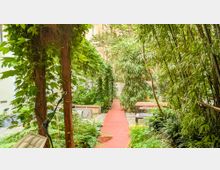 Ein grüner Innenhof mit dichtem Pflanzenbewuchs, darunter Bambus und Kletterpflanzen, sowie ein schmaler, roter Steg, der durch die Szene führt. Im Hintergrund stehen Holzbänke und Tische, die von Vegetation umgeben sind.