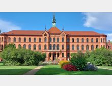 Backsteingebäude im neogotischen Stil mit symmetrischer Fassade, zahlreichen Rundbogenfenstern und einem kleinen Uhrturm in der Mitte; im Vordergrund ein gepflegter Garten mit Blumenbeeten, Büschen und Wegen.