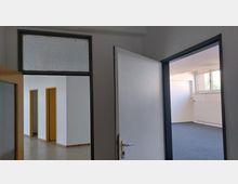 Blick in einen leeren Büro- oder Arbeitsraum mit weiß gestrichenen Wänden. Zu sehen sind geöffnete Türen, ein Raum mit grauem Teppichboden, ein Heizkörper unter großen Fenstern sowie ein angrenzender Bereich mit gelben Türrahmen und Betonboden.