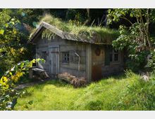Holzhütte mit begrüntem Dach in einer natürlichen Umgebung, umgeben von Bäumen und Sträuchern. Die kleine Hütte hat schmale Fenster, eine Holztür und eine Bank davor, mit Gras und Pflanzen, die sie umgeben.