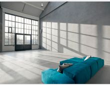 Großer, lichtdurchfluteter Raum mit hohen Decken, einer grauen Wand und großen Metallrahmen-Fenstern, die Ausblick auf kahle Bäume und Gebäude bieten. Im Vordergrund steht ein modernes, modulartiges, türkisfarbenes Sofa mit Kissen auf einem grauen Betonboden.