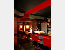 Eine stilvolle Bar mit dunkelroten und schwarzen Akzenten, gepolsterten Barhockern und geometrischen Mustern an der Theke. Im Hintergrund befinden sich Holztische und Stühle, umgeben von Tapeten mit dekorativen Mustern und dezenter Beleuchtung.