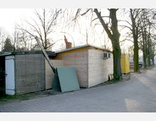 Eine Reihe von provisorischen Holzhütten mit schlichten Bauweisen, flankiert von Bäumen ohne Laub. Im Hintergrund steht ein gelber Container, und daneben sind weitere temporäre Bauten sichtbar, auf einem gepflasterten Weg gelegen.