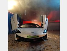 Ein weißer Sportwagen mit geöffneten Flügeltüren steht auf sandigem Boden vor einer Kulisse aus beleuchteten Schiffscontainern, umgeben von leichtem Nebel.