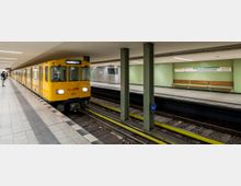 Gelbe U-Bahn fährt in eine unterirdische Haltestelle mit grün gefliesten Wänden ein, benannt "Kurfürstendamm". Auf der gegenüberliegenden Plattform stehen Holzbänke, und das Gleis ist von Säulen und Beleuchtung umgeben.