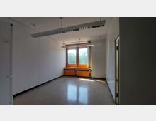 Ein leerer Raum mit weißen Wänden und einem Boden aus hellen Fliesen. Zwei große Fenster mit orangefarbenen Rahmen lassen Tageslicht herein, an der Decke hängen weiße, lineare Leuchtstofflampen.